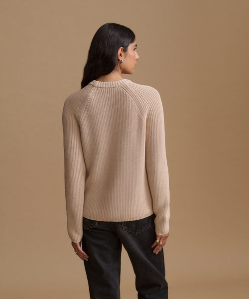 A woman shown from the back wearing a ribbed knit sweater in a light beige color, paired with dark jeans, against a solid tan studio background.