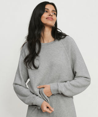 A woman with long dark hair wearing the Saturday Sweatshirt in light gray stands against a plain white background, gazing to the side with a relaxed expression and holding the hem of her sleeve.