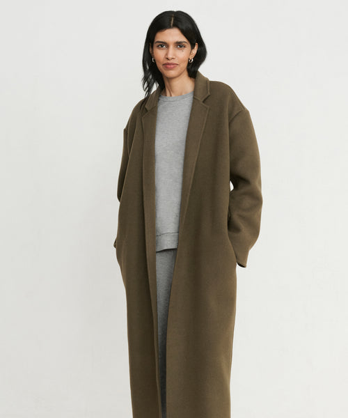 A woman with long dark hair stands against a white background, hands in her coat pockets, wearing a long olive-brown coat over the Saturday Sweatshirt in light gray French terry and matching pants, looking at the camera with a soft smile.