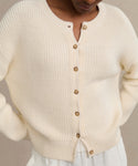 Wearing the Cotton Hayes Crewneck Cardigan—a cream, ribbed summer knit with brown buttons—over a white skirt or pants, this person stands with hands on hips, head partially out of frame.