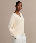 A woman stands against a plain beige background, hands in her pockets, wearing the cream Cotton Hayes Crewneck Cardigan with brown buttons and white pants, looking at the camera.
