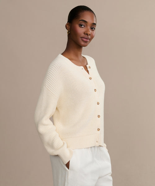A woman stands against a plain beige background, hands in her pockets, wearing the cream Cotton Hayes Crewneck Cardigan with brown buttons and white pants, looking at the camera.