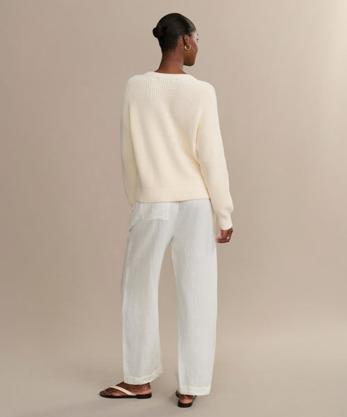 A woman with a neat bun faces away, wearing the Cotton Hayes Crewneck Cardigan in off-white, loose white pants, and black sandals against a beige background.