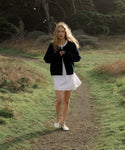 Woman walking on a path in a natural setting wearing a navy cardigan, white dress and white shoes.
