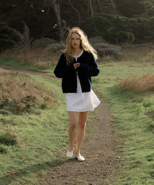 Woman walking on a path in a natural setting wearing a navy cardigan, white dress and white shoes.
