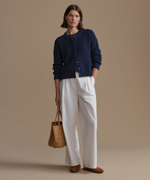 Woman wearing a navy cardigan, white pants, and brown shoes, holding a woven bag against a beige background.