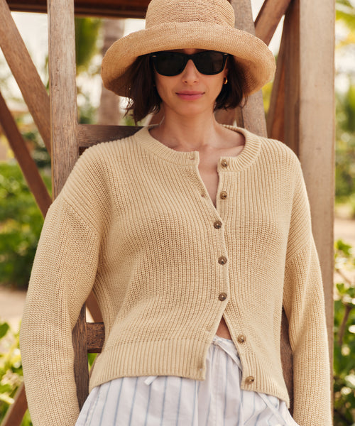 Woman wearing a beige knit cardigan, sunglasses, and a straw hat outdoors.