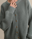 Someone wears the oversized Cotton Hayes Crewneck Cardigan in grayish-green cotton with brown buttons and a ribbed texture. Their ring-adorned hand is raised near their face, partially covering their mouth.