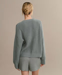 With their back to the camera, a person with blonde hair in a low bun models an oversized gray Cotton Hayes Crewneck Cardigan and matching shorts—a breezy summer knit—set against a plain beige background.