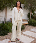 A woman stands outdoors on a stone path, wearing Jones Trouser cream wide-leg pants with black vertical stripes and a loose white cardigan. She pairs the look with brown sandals, poses with one hand near her face, and is surrounded by greenery.