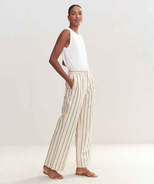 A woman poses sideways with her hands in the pockets of cream, wide-leg Jones Trouser pants featuring black stripes. She wears them with a sleeveless white top and brown sandals against a light background.