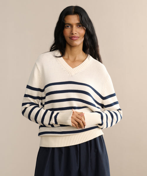 Woman with long dark hair wears the Cotton Kate V-Neck, a cream-striped cotton sweater, paired with a navy skirt against a beige backdrop—smiling with hands clasped for an ideal spring knit look.