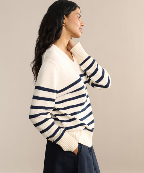 A woman with long dark hair, wearing the Cotton Kate V-Neck striped sweater and navy pants, stands in profile with her hand touching her chin, smiling slightly against a plain beige background.