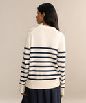 A woman with long dark hair, seen from behind, wears the Cotton Kate V-Neck—a cream and navy striped spring knit sweater—with a dark skirt against a plain neutral background.