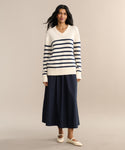 A woman with long dark hair and a relaxed expression wears the Cotton Kate V-Neck in navy and cream stripes, paired with a long navy skirt and cream flats—a perfect spring knitwear look against a neutral backdrop.