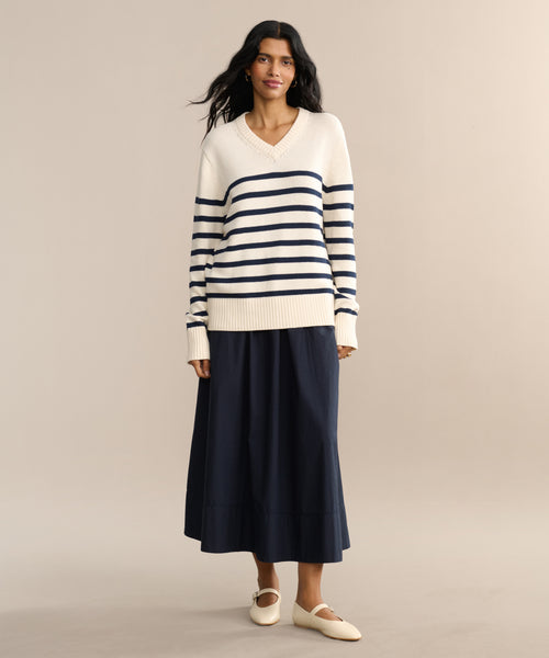 A woman with long dark hair and a relaxed expression wears the Cotton Kate V-Neck in navy and cream stripes, paired with a long navy skirt and cream flats—a perfect spring knitwear look against a neutral backdrop.