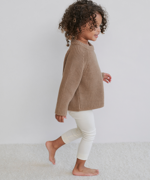 A young child with curly hair walks barefoot on a light carpet, wearing the Kids Cotton Fisherman sweater and white ribbed leggings, set against a plain light background.