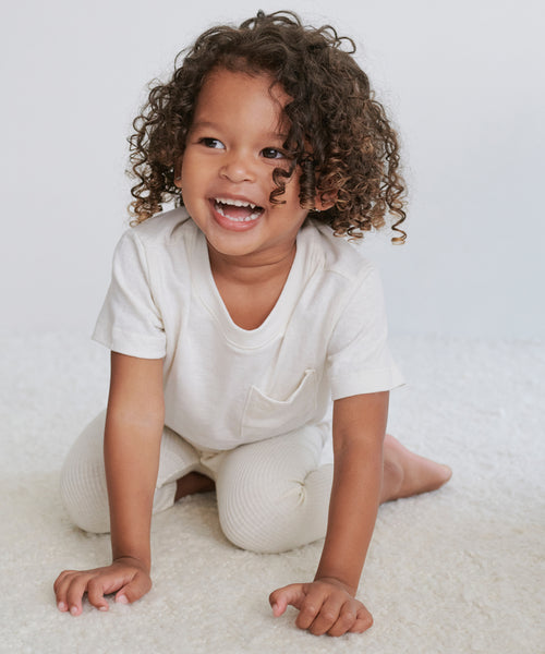 A young child with curly hair, wearing a Kids Vintage Pocket Tee and matching pants, kneels and smiles brightly on a soft white surface against a plain background.