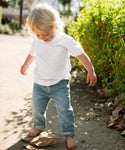 A young blonde child, barefoot in rolled-up jeans and an ivory Kids Vintage Pocket Tee, stands on a dirt path outdoors gazing down at a crumpled brown paper bag, with greenery in the background.