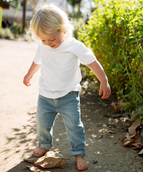 A young blonde child, barefoot in rolled-up jeans and an ivory Kids Vintage Pocket Tee, stands on a dirt path outdoors gazing down at a crumpled brown paper bag, with greenery in the background.