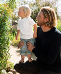 A man with long blond hair kneels outdoors, holding a barefoot toddler in jeans and an ivory Kids Vintage Pocket Tee, as they look at greenery in a sunlit garden.