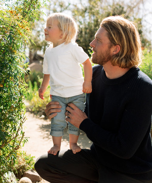A man with long blond hair kneels outdoors, holding a barefoot toddler in jeans and an ivory Kids Vintage Pocket Tee, as they look at greenery in a sunlit garden.