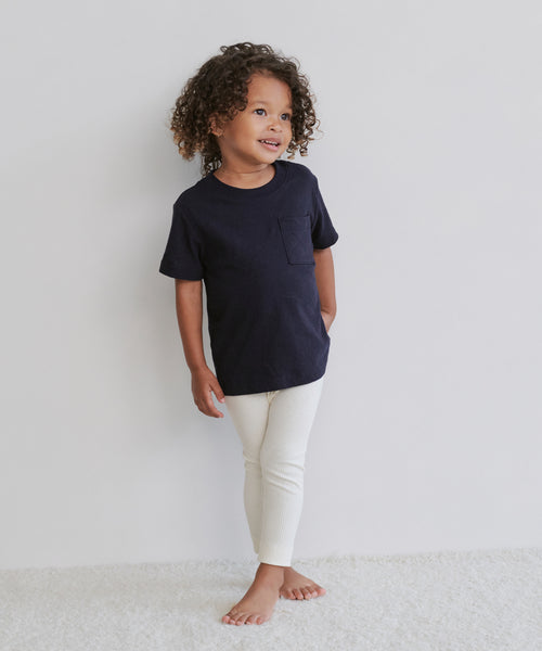 A young child with curly hair stands barefoot against a white wall, smiling to the side while wearing the Kids Vintage Pocket Tee—a soft, 100% cotton shirt featuring a classic pocket for vintage style.