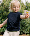 A young child with blonde hair, wearing the Kids Vintage Pocket Tee and light-colored pants, stands outdoors holding a small yellow flower in front of green bushes.