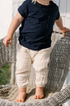 A small child stands barefoot on a woven chair, wearing cream-colored pants and a navy Kids Vintage Pocket Tee. The child’s face is partially out of frame, and sunlight highlights the classic 100% cotton tee.