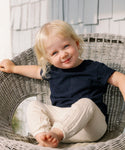 A young child with blond hair smiles at the camera while sitting in a wicker chair outdoors, wearing the Kids Vintage Pocket Tee in navy blue along with light-colored pants.