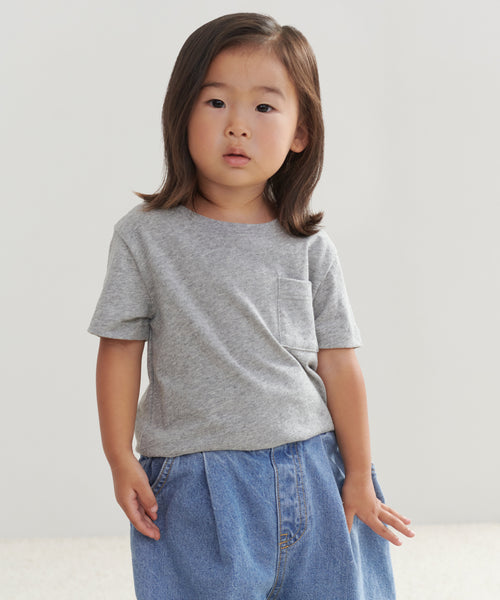A young child with straight, shoulder-length brown hair stands against a plain light background, wearing the Kids Vintage Pocket Tee in grey and blue jeans, with hands resting at their sides.