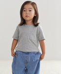 A young child with long brown hair wears the Kids Vintage Pocket Tee and loose blue jeans, standing with hands in pockets against a plain, light background.
