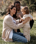 A smiling woman kneels on grass, gently holding a toddler in a Kids Vintage Pocket Tee, while a smiling man crouches behind them outdoors on a sunny day. All three appear happy, sharing a tender family moment.