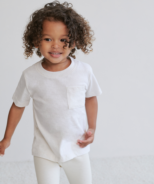A young child with curly hair smiles while standing in an ivory Kids Vintage Pocket Tee—100% cotton with a classic chest pocket—paired with light pants against a plain, light background.