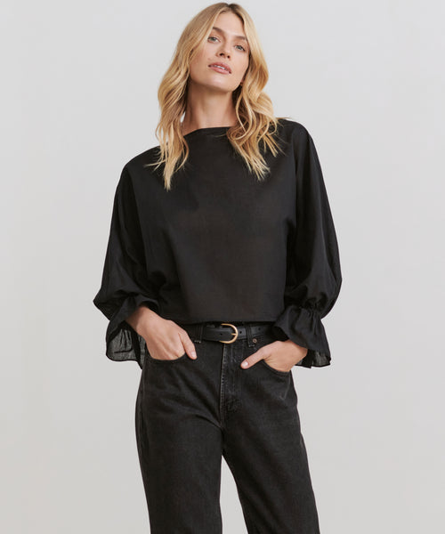 A woman with long blonde hair models the Cape Blouse, a loose-fitting top with ruffled cuffs, paired with black high-waisted jeans, standing with one hand in her pocket against a plain light background.