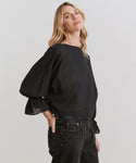 A woman with long blonde hair wears a loose-fitting sheer Cape Blouse with voluminous sleeves and black jeans, standing against a plain light background, gazing slightly upward.