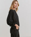 A woman with blonde wavy hair stands in profile against a plain light background, wearing a black Cape Blouse with puffed sleeves and black jeans, one hand in her pocket.