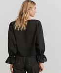 A woman with blonde hair models the Cape Blouse—a loose-fitting, boxy black top with ruffled cuffs—paired with black jeans, standing with her back to the camera against a plain light background.