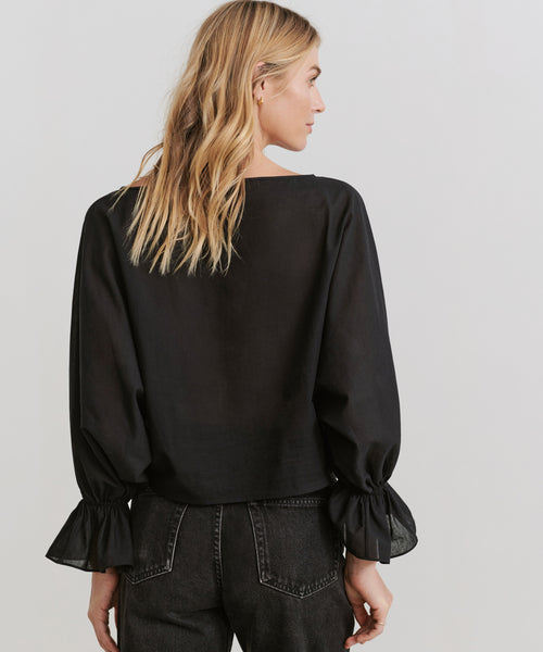 A woman with blonde hair models the Cape Blouse—a loose-fitting, boxy black top with ruffled cuffs—paired with black jeans, standing with her back to the camera against a plain light background.