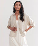 A woman with long dark hair wears the Georgia Jacket, a light beige cotton-linen layer, over a white dress and stands with one hand in her pocket against a plain light background.