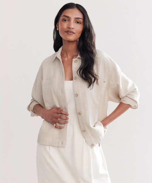 A woman with long dark hair wears the Georgia Jacket, a light beige cotton-linen layer, over a white dress and stands with one hand in her pocket against a plain light background.