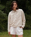 A woman with shoulder-length brown hair wears the Georgia Jacket, a light beige cotton-linen piece with large front pockets, paired with white shorts while standing outside in front of green foliage.