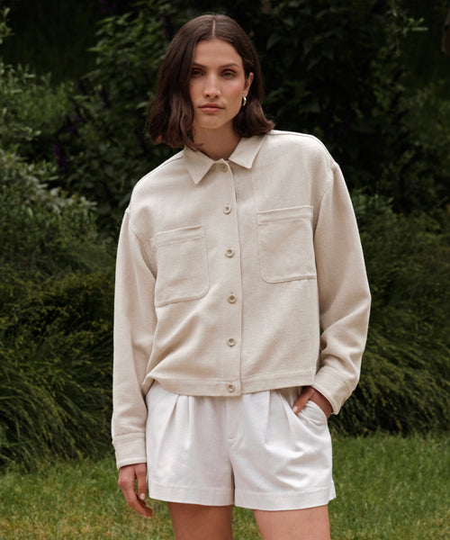A woman with shoulder-length brown hair wears the Georgia Jacket, a light beige cotton-linen piece with large front pockets, paired with white shorts while standing outside in front of green foliage.