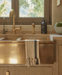 A brass farmhouse sink with a gold faucet and beige countertop features the Striped Hand Towel hanging over the sink. Nearby, a soap dispenser and a bowl of artichokes sit on the counter by a window overlooking greenery.
