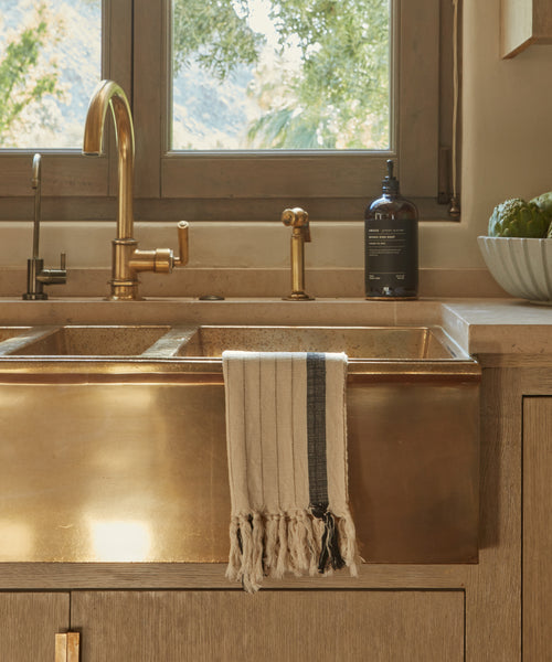 A brass farmhouse sink with a gold faucet and beige countertop features the Striped Hand Towel hanging over the sink. Nearby, a soap dispenser and a bowl of artichokes sit on the counter by a window overlooking greenery.