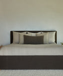 A neatly made bed with a dark brown headboard and frame, light beige bedding, and several pillows—including the Luna Lumbar Pillow—in beige and dark brown shades, set against a plain light-colored wall.