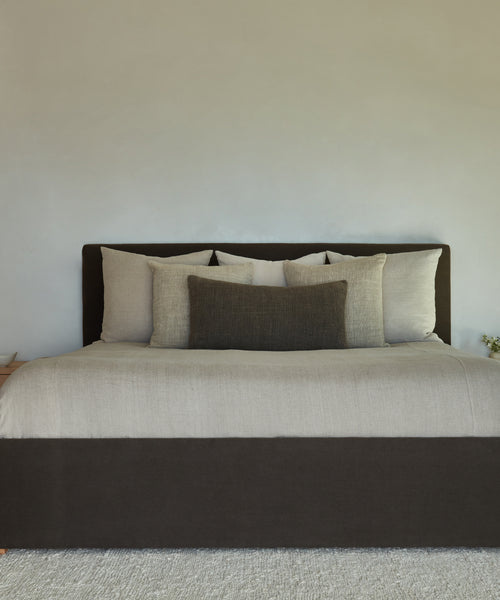 A neatly made bed with a dark brown headboard and frame, light beige bedding, and several pillows—including the Luna Lumbar Pillow—in beige and dark brown shades, set against a plain light-colored wall.
