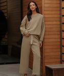 A woman in front of wooden paneling wears the Marina Sweatpant—a loose, beige, wide-leg, 100% cotton loungewear piece with a drawstring waist—paired with a matching sweatshirt, looking calmly at the camera.