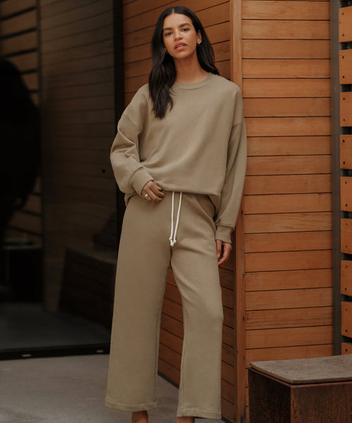 A woman in front of wooden paneling wears the Marina Sweatpant—a loose, beige, wide-leg, 100% cotton loungewear piece with a drawstring waist—paired with a matching sweatshirt, looking calmly at the camera.