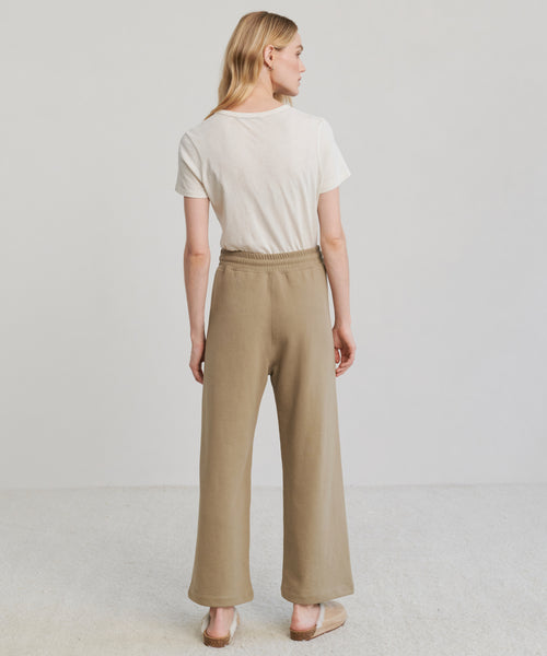 A woman with light hair faces away from the camera, wearing a white tee and the Marina Sweatpant—100% cotton, high-waisted wide-leg sweatpants—in a light, neutral setting for the perfect modern loungewear look.